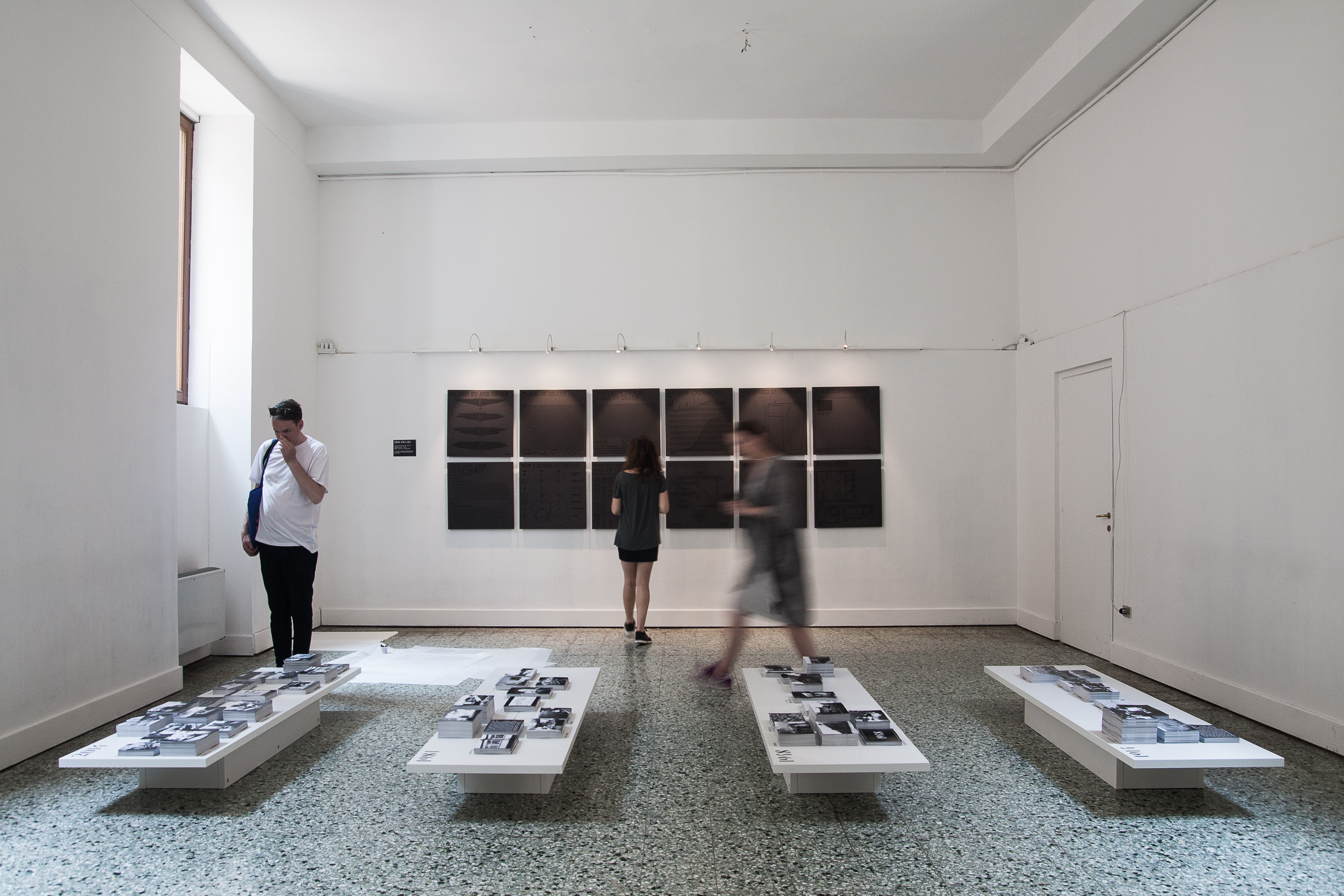 The final room in the Death in Venice exhibition. Panels on the back wall are infographics on death-related statistics, printed as reliefs so that visitors could make souvenir newsprint-and graphite-rubbings. Tables in the foreground hold postcards with information about death-related spaces, covering 1914, 1948, 1981 and 2014. Photo: A. Molenda