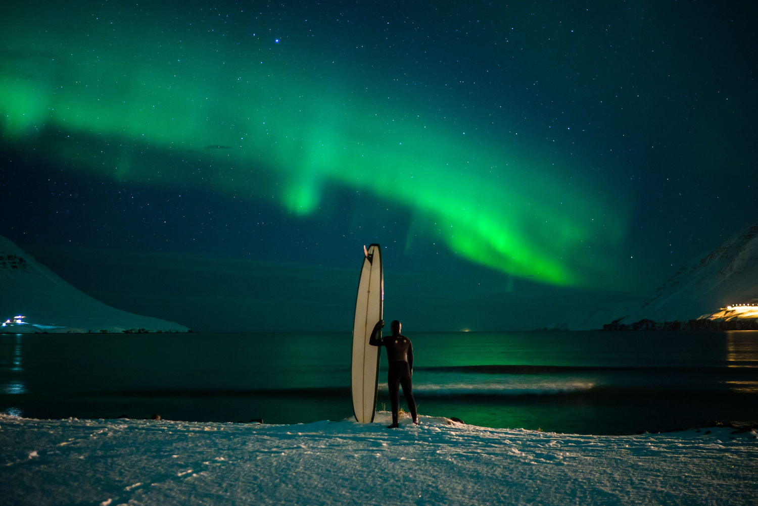 man standing on snowy coast with surf board looking out over the water with northern lights above